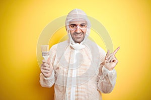 Young man wearing painter equipment and holding painting brush over isolated yellow background very happy pointing with hand and