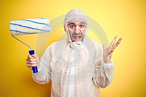 Young man wearing painter equipment and holding painting roller over isolated yellow background very happy and excited, winner