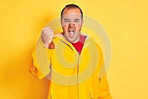 Young man wearing rain coat standing over isolated yellow background angry and mad raising fist frustrated and furious while