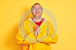 Young man wearing rain coat standing over isolated yellow background begging and praying with hands together with hope expression