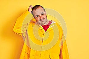 Young man wearing rain coat standing over isolated yellow background confuse and wonder about question