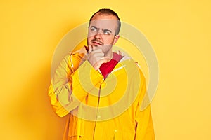 Young man wearing rain coat standing over isolated yellow background with hand on chin thinking about question, pensive expression