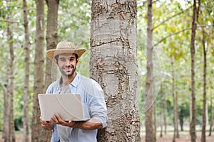 A young man wearing a straw hat leans against a tree with a laptop in his hand.