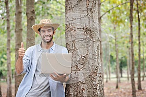 A young man wearing a straw hat leans against a tree with a laptop in his hand.