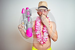 Young man wearing summer hat and hawaiian lei flowers holding water gun over isolated background cover mouth with hand shocked