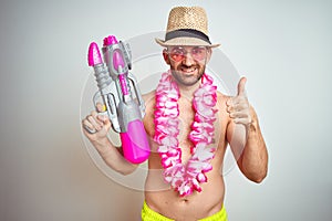 Young man wearing summer hat and hawaiian lei flowers holding water gun over isolated background happy with big smile doing ok