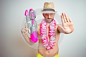 Young man wearing summer hat and hawaiian lei flowers holding water gun over isolated background with open hand doing stop sign
