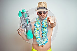 Young man wearing summer hat and hawaiian lei flowers holding water gun over isolated background pointing with finger to the