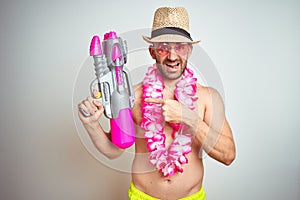 Young man wearing summer hat and hawaiian lei flowers holding water gun over isolated background very happy pointing with hand and