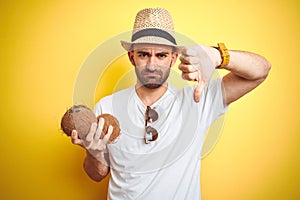 Young man wearing summer hat and holding coconut fruit over yellow background with angry face, negative sign showing dislike with