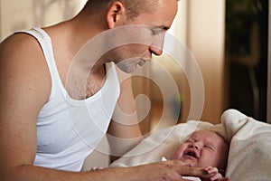 Young man wearing only vest top, playing with his baby son, trying to calm down, screaming kid as he holds him on hands