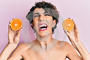 Young man wearing woman make up holding fresh orange angry and mad screaming frustrated and furious, shouting with anger looking