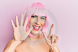 Young man wearing woman make up wearing pink wig showing and pointing up with fingers number seven while smiling confident and