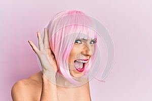 Young man wearing woman make up wearing pink wig smiling with hand over ear listening an hearing to rumor or gossip