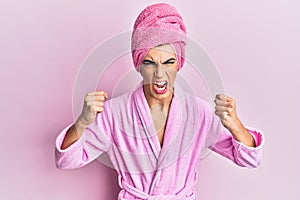 Young man wearing woman make up wearing shower towel on head and bathrobe angry and mad raising fists frustrated and furious while