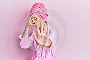 Young man wearing woman make up wearing shower towel on head and bathrobe covering eyes with hands and doing stop gesture with sad