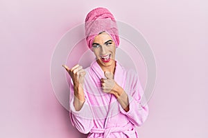 Young man wearing woman make up wearing shower towel on head and bathrobe pointing to the back behind with hand and thumbs up,