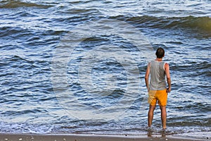 young man wearing yellow swimming shorts and a gray t-shirt stands on the shore and looks at the ocean