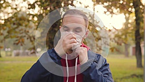 A young man wears a medical disposable protective mask and looks into the camera on the background of an autumn park