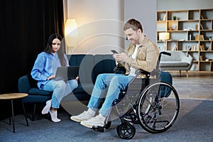 Young man in wheelchair holding mobile phone in hands and his wife using laptop at home