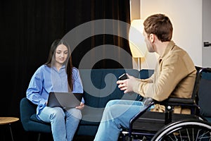 Young man in wheelchair holding mobile phone in hands and his wife using laptop at home