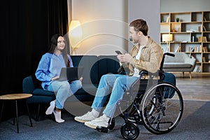Young man in wheelchair holding mobile phone in hands and his wife using laptop at home