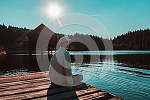 Young man in white cap sit on small wooden wharf on pond with wooden building and blue sky