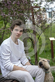 A young man in white clothes sits on a bench in a spring park with his dog.