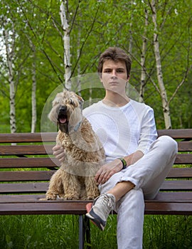 A young man in white clothes sits on a bench in a spring park with his dog.