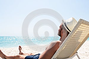 Young man in white hat relaxing in deck chair on beautiful sandy beach