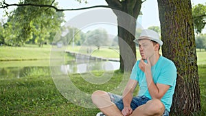 Young man in white hat sits thoughtfully under a tree in the park, tracking the camera