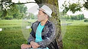 Young man in a white hat sits on a tree, looks into the distance, tracking camera