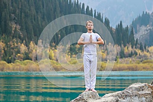 A young man in white practices yoga in the mountains. Pose Samasthiti namaskar