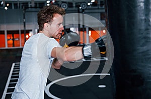 Young man in white shirt and boxing protective gloves doing exercises in gym with pushing bag