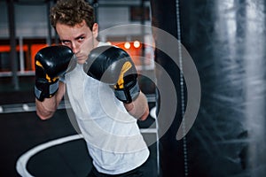 Young man in white shirt and boxing protective gloves doing exercises in gym with pushing bag
