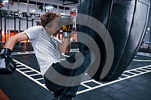 Young man in white shirt and boxing protective gloves doing exercises in gym with pushing bag