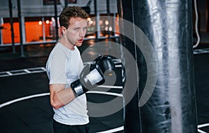 Young man in white shirt and boxing protective gloves doing exercises in gym with pushing bag