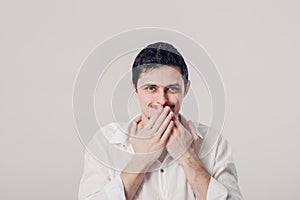 Young man in white shirt covers his mouth with his hands on gray