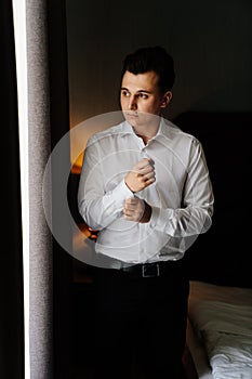 young man in a white shirt fastens cufflinks on sleeves near the window