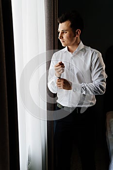 young man in a white shirt fastens cufflinks on sleeves near the window