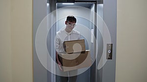 Young man in white shirt getting out of the elevator with cardboard boxes moving in to new appartment