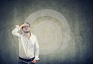 Young man in white shirt and glasses having problems and looking up in challenge on gray wall background