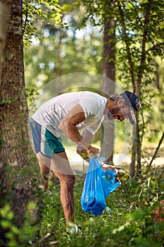 Young man in white shirt in a green spring forest in a sunny weather, holding blue plastic bag collecting trash. Volunteers in