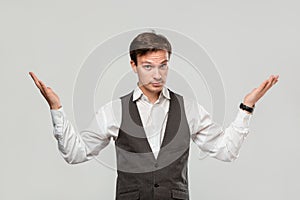 Young man in a white shirt and grey vest shrugging his shoulders expressing emotion of confusion.