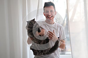 Young man in the white shirt and pajama is smiling holding fluffy cat which wants to run away