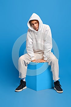 Young man in white sweatshirt with hood and white sweatpants sits on wooden cube over blue background