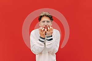 Young man in a white sweatshirt is worried on the background of a red wall, looking at the camera and biting his nails from nerves