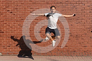 Young man in white t-shirt jump with crosed hands, spread his arms wide and looking at the camera, red brick wall on the