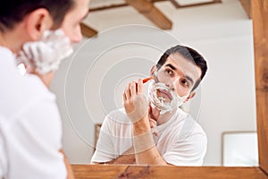 Young man in white T-shirt shaves while standing near mirror in bath