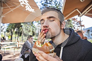 Young man wipes his mouth with his right hand at satisfaction, his eyes are closed. He is holding bitten hamburger at his left